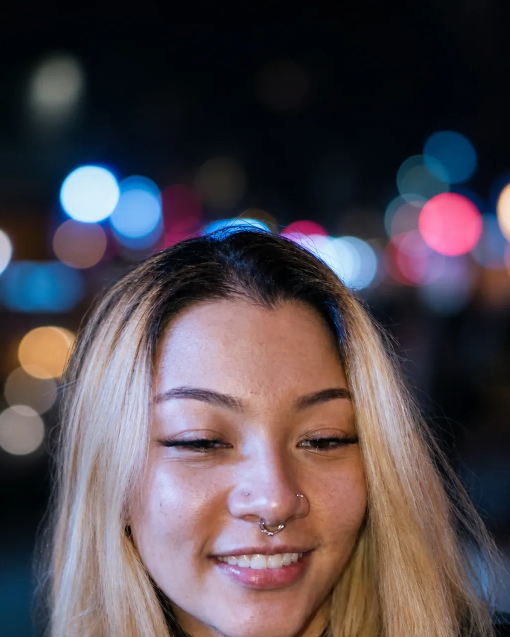 The same woman lit cleanly at night with city neon behind her as coloured bokeh, piercings and hair unchanged