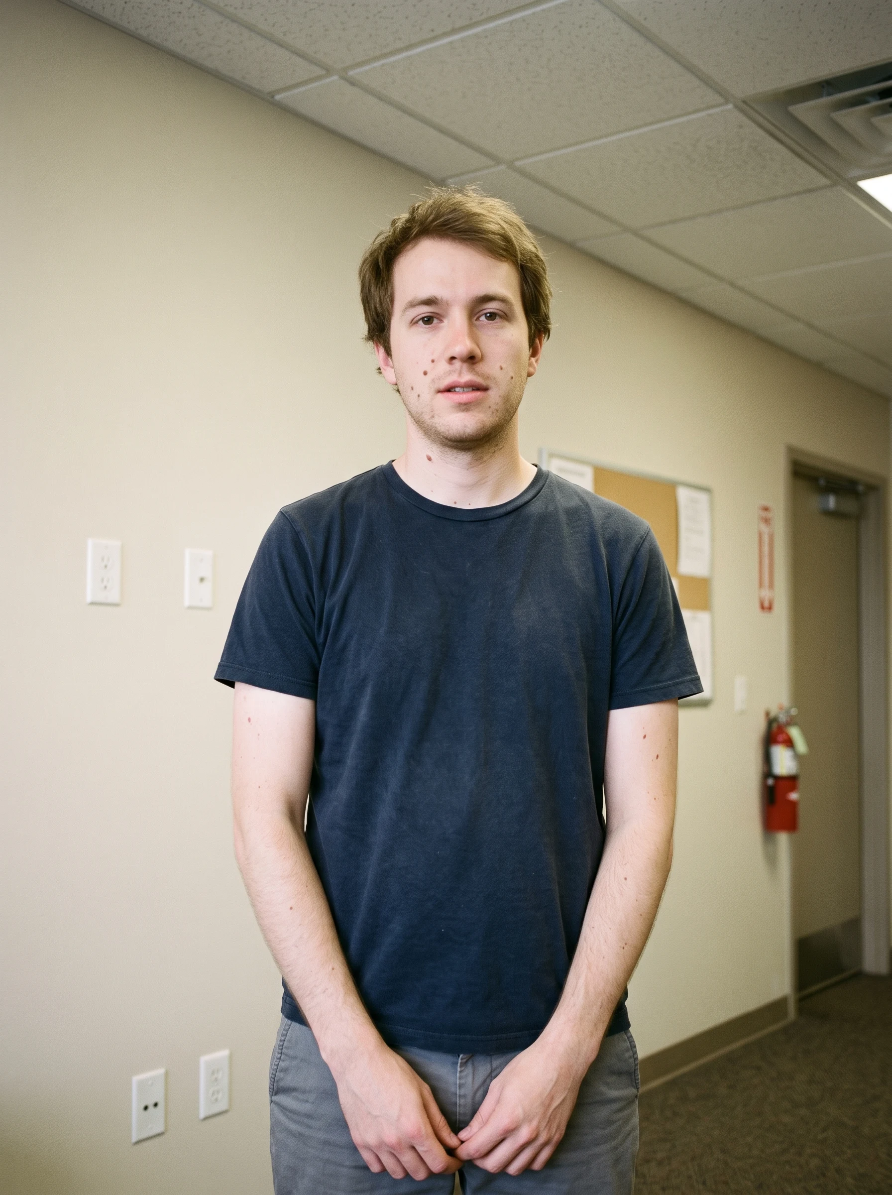 Man standing in a beige office corridor under flat overhead fluorescent light, before AI relighting