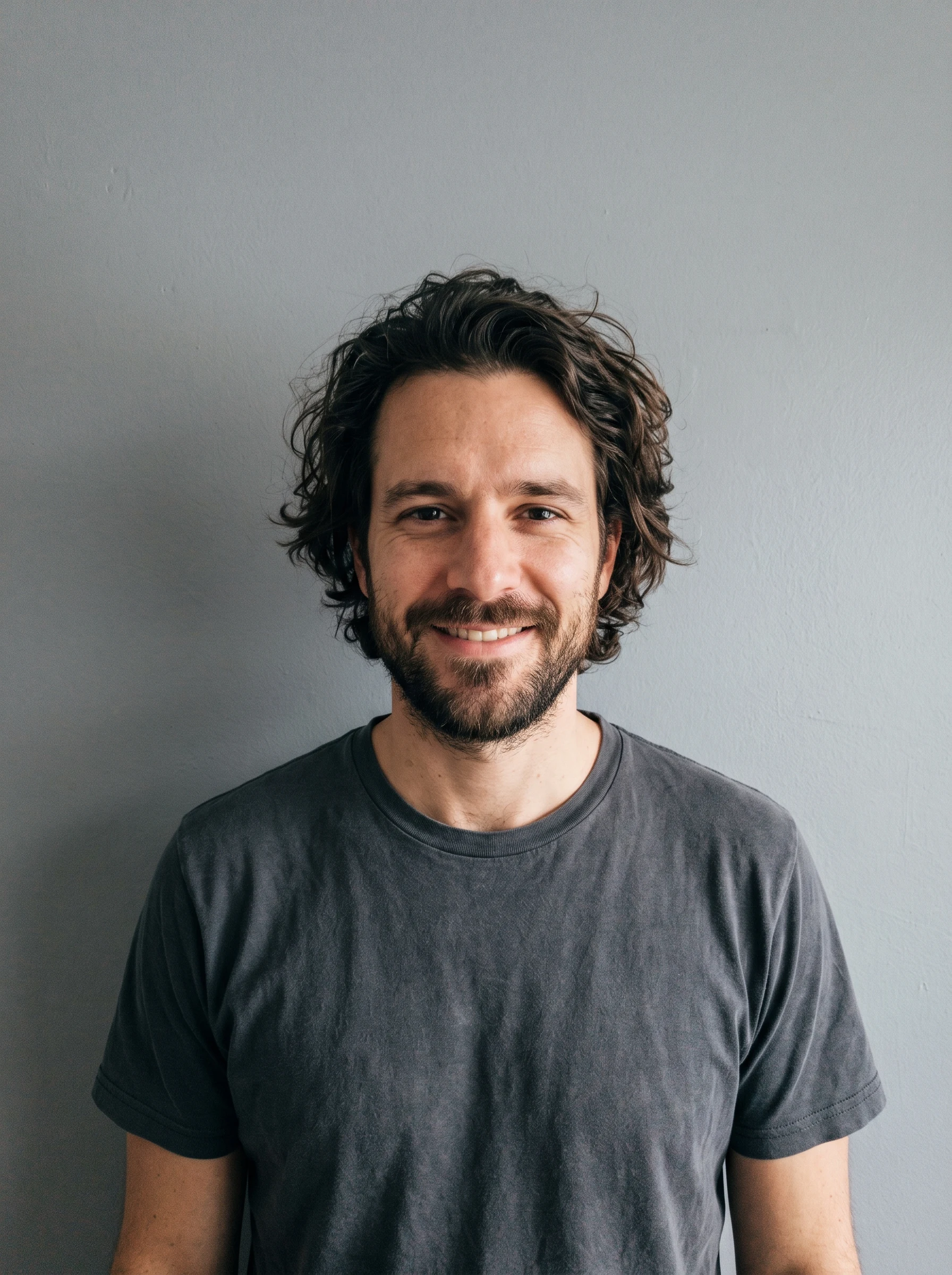 Man with medium-length messy curly brown hair and a short beard against a grey wall