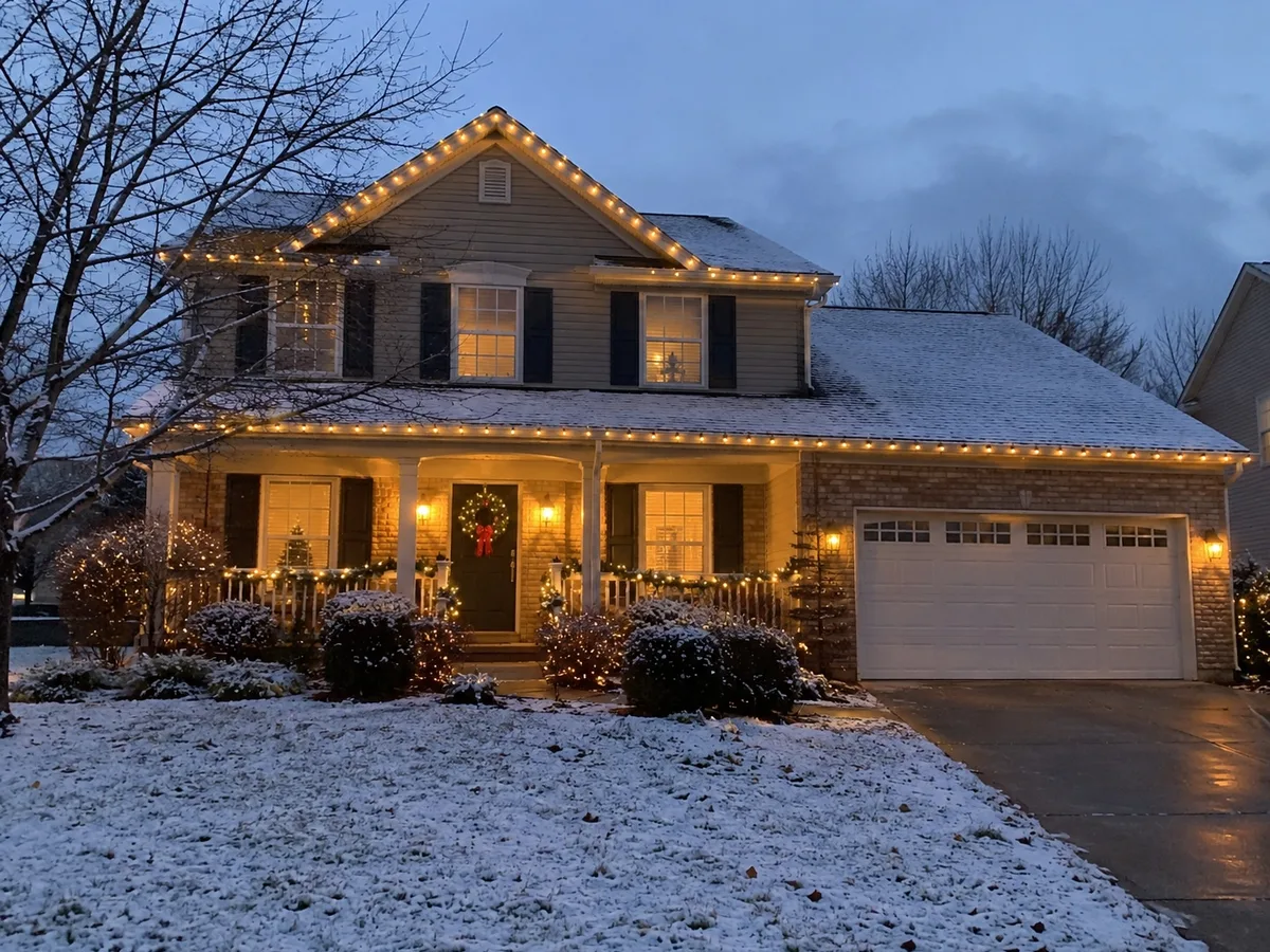 Suburban house at dusk with Christmas lights along the roof and snow on the lawn