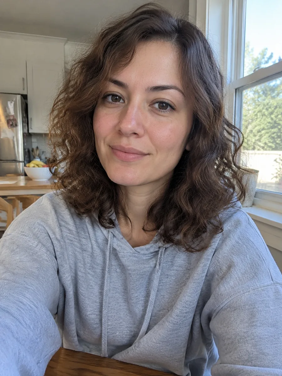 Casual kitchen selfie of a woman with curly brown hair in a grey hoodie
